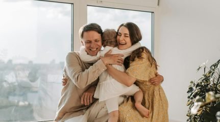A father and mother hugging their small daughter beside a window