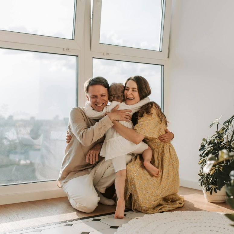 A father and mother hugging their small daughter beside a window