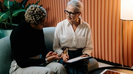 Two women talking over papers on a sofa in front of an orange panelled wall