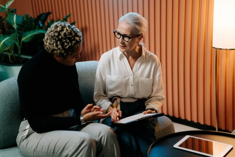 Two women talking over papers on a sofa in front of an orange panelled wall