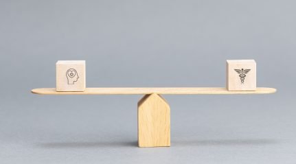 A wooden balance scale weighing a block marked with a head-and-brain icon against a block marked with a medical caduceus