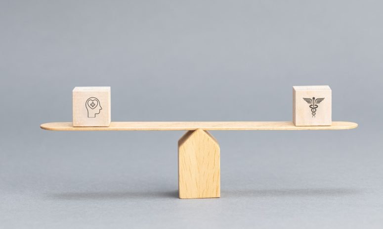 A wooden balance scale weighing a block marked with a head-and-brain icon against a block marked with a medical caduceus