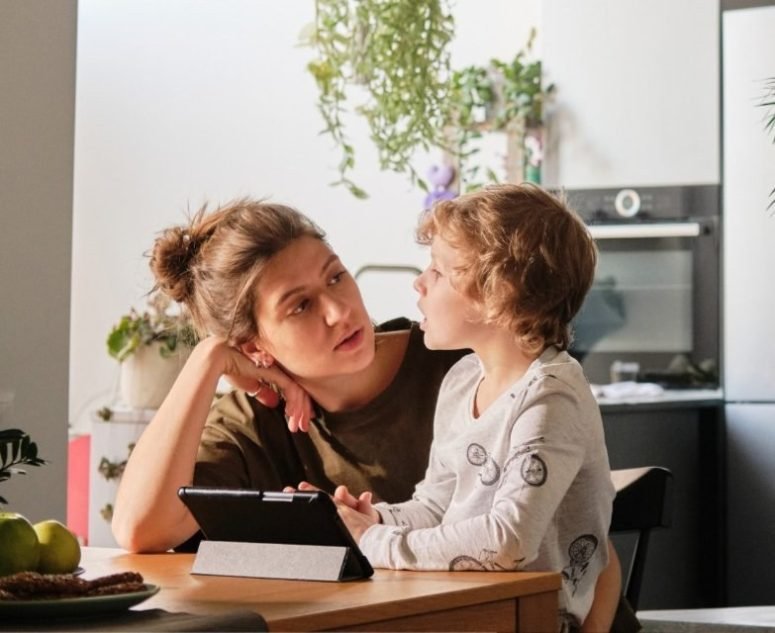 A mother and her son looking at each other across a kitchen table with a tablet