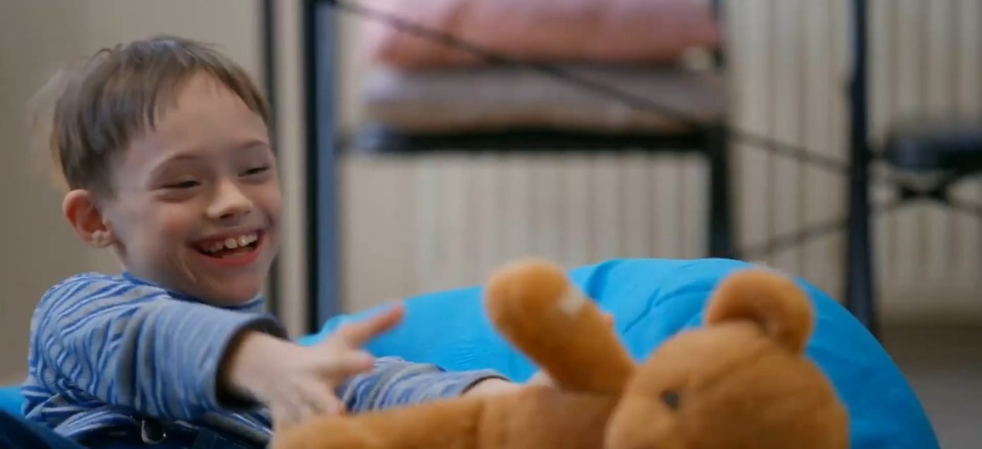 A smiling boy in a striped shirt reaching for a teddy bear on a blue beanbag — poster frame for the page video