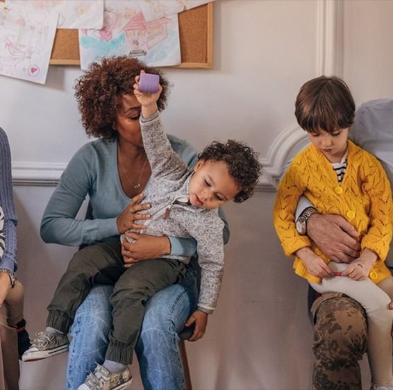 Three parents sitting in a row in a waiting room, each with a young child on their lap