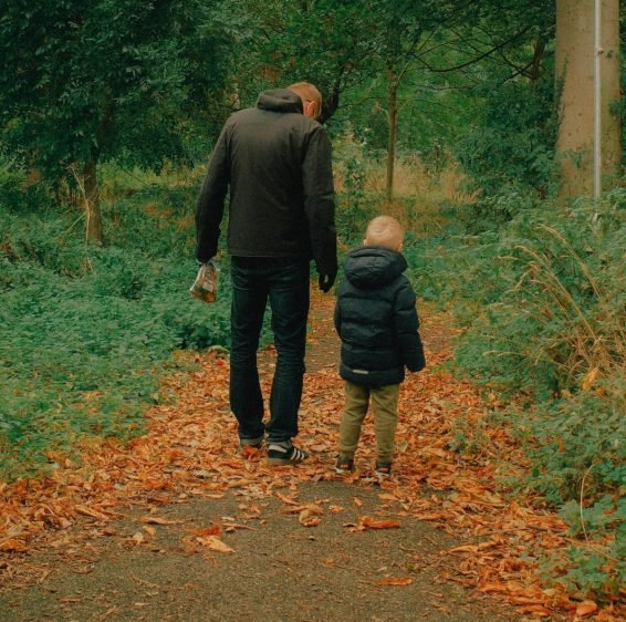 A father and his young son walking side by side along a leaf-covered woodland path, seen from behind