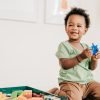A laughing toddler holding a blue toy on the floor beside a box of wooden blocks