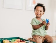 A laughing toddler holding a blue toy on the floor beside a box of wooden blocks