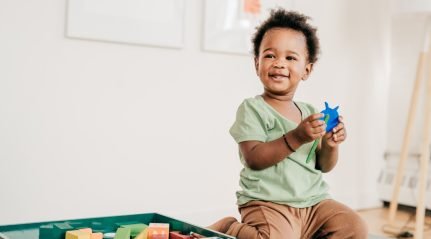 A laughing toddler holding a blue toy on the floor beside a box of wooden blocks