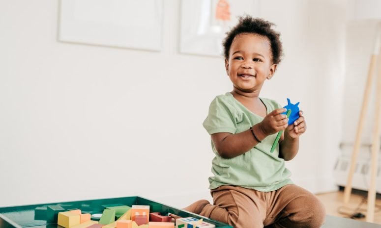 A laughing toddler holding a blue toy on the floor beside a box of wooden blocks