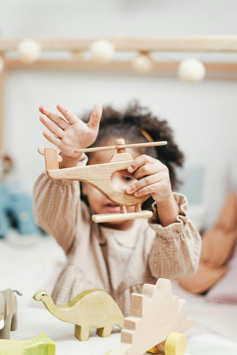 A child holding a wooden toy aeroplane up to one eye, with a wooden dinosaur and blocks in front