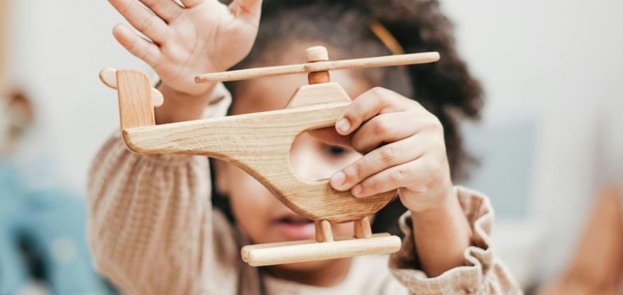A child holding a wooden toy aeroplane up to one eye, with a wooden dinosaur and blocks in front