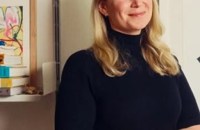 A woman with long blonde hair standing with her arms folded beside a bookshelf, smiling
