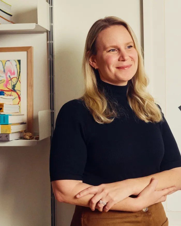 A woman with long blonde hair standing with her arms folded beside a bookshelf, smiling