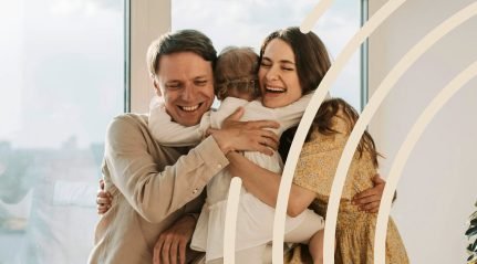 A father and mother hugging their small daughter beside a window