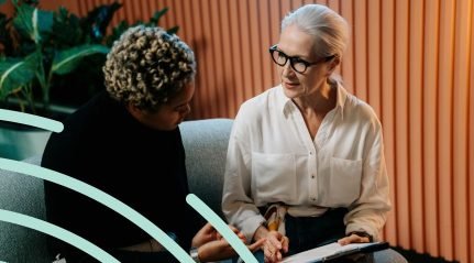 Two women talking over papers on a sofa in front of an orange panelled wall