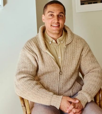 A smiling young man in a knitted cardigan sitting in a chair with his hands folded