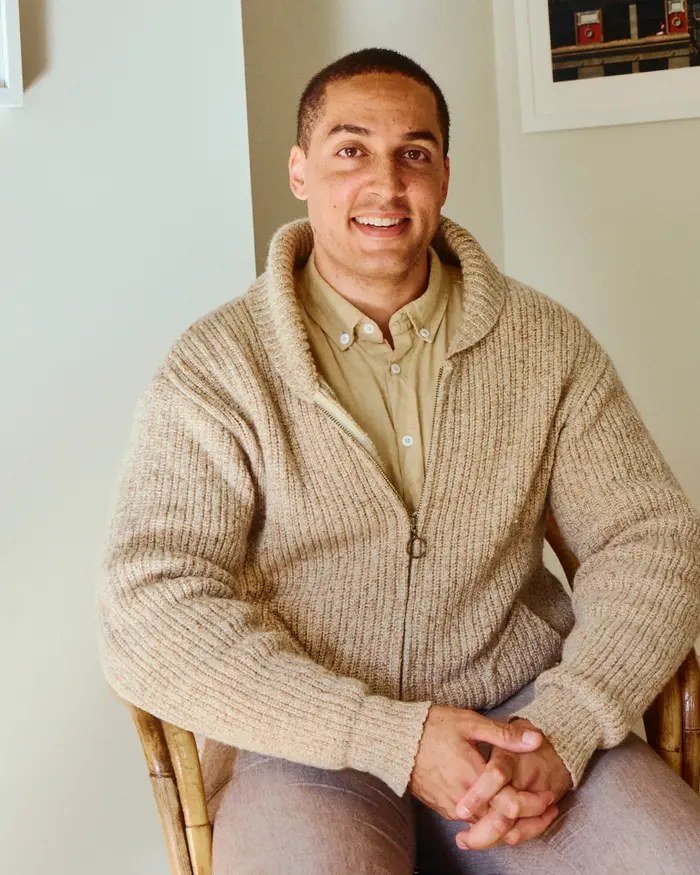 A smiling young man in a knitted cardigan sitting in a chair with his hands folded