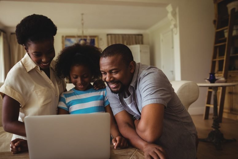 A mother, father and their young daughter smiling at a laptop on the sofa
