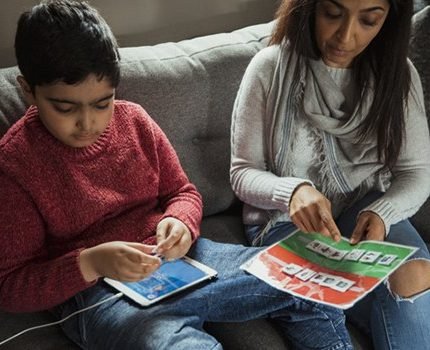 A boy with a tablet and a woman reading a picture book together on a sofa