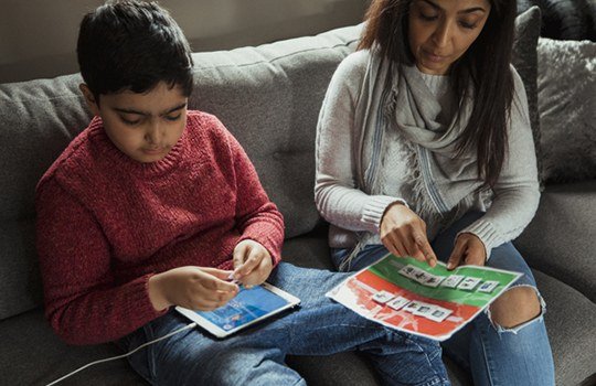 A boy with a tablet and a woman reading a picture book together on a sofa