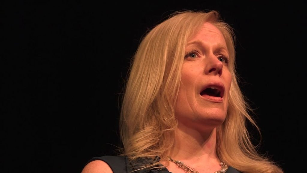 A woman with long blonde hair speaking on a dark stage